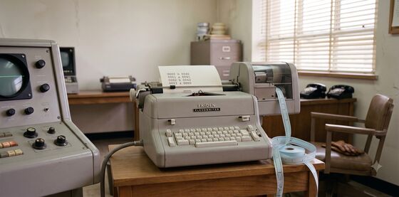 A Friden Flexowriter on a side table beside the LGP-30 console, with punched paper tape spilling off the table